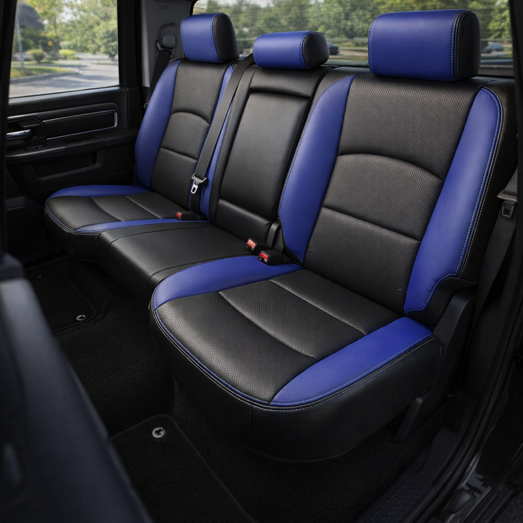 Rear in-cabin view of a 2012–2018 Ram 1500/2500/3500 with an installed Leatherteck leather kit in black with Cobalt Blue panels, perforated black inserts, and Cobalt stitching. The image shows the upgraded rear bench seat with three headrests, black perforated seat and backrest inserts, Cobalt Blue side bolsters and seat cushion accent panels, Cobalt Blue headrest faces, matching contrast stitching, seatbelts and red buckles, rear door panel, rear window, and dark carpeted floor inside the truck cabin.