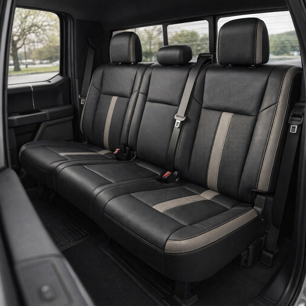  Rear in-cabin view of a 2015–2019 Ford F-150 with an installed Leatherteck leather upholstery kit in black with Stone accent panels and perforated black inserts. The image shows the upgraded rear bench seat with three headrests, black leather bolsters, perforated black seat and backrest insert sections, vertical Stone accent stripes, Stone side accent panels, contrast stitching, seatbelts and buckles, rear door panel, rear window, and black all-weather floor mat inside the truck cabin.