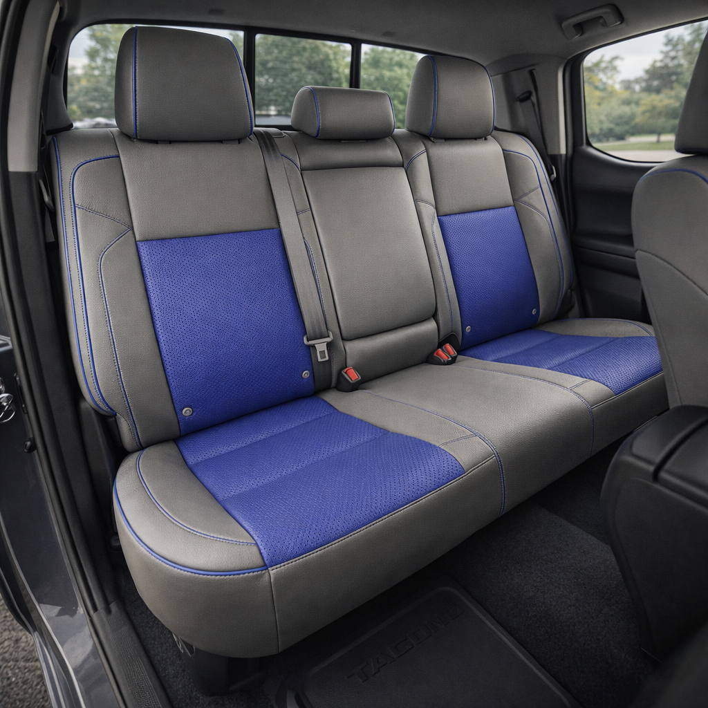 Rear in-cabin view of a 2016–2023 Toyota Tacoma with an installed Leatherteck leather upholstery kit in light grey featuring cobalt blue perforated center inserts, cobalt blue seat cushion inserts, and cobalt blue contrast stitching/piping, shown through the open rear door with the rear bench seat, three headrests, seatbelts/buckles, front seatback, rear door panel, and black Tacoma floor mat visible.