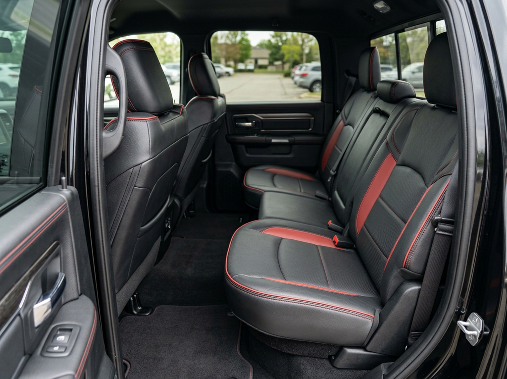  Rear in-cabin view of a 2019–2026 RAM 2500 showing a Leatherteck leather seat upholstery kit in black with perforated body inserts, medium red accent panels, medium red contrast stitching, and medium red edge piping, photographed through the open rear door with the rear bench seat, headrests, seatbelts/buckles, rear door panel, and RAM-branded floor liner visible.

